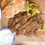 Strimlor av kebab ligger på en skärbräda med pitabröd, vitlökssås och andra tillbehör runtom.