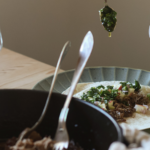 En hand serverar ramslöksalsa på ett tortillabröd med tacos. I förgrunden står en gjutjärnsgryta med kött i - pulled lammstek.